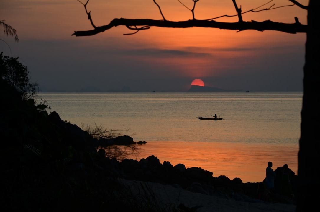 Experience Thailand's Vibrant Culture from Bangkok to Koh Phangan 18 Thailand temple bangkok Sunset over the gulf of thailand on Koh Phangan with a little boat on the water and silhouettes of people stones plants and a tree.
